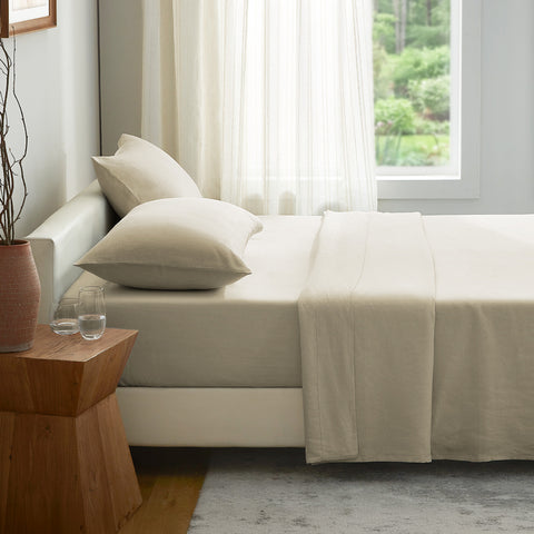 Beige linen bed sheet and pillows on a bed in a room with a wooden side table and window.