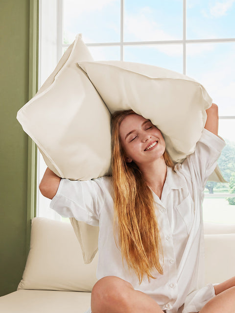 Woman holding two large pillows above her head in a bright room with a window.